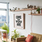 Modern living room with a 'Come as You Are' framed quote, plants, and a sofa.