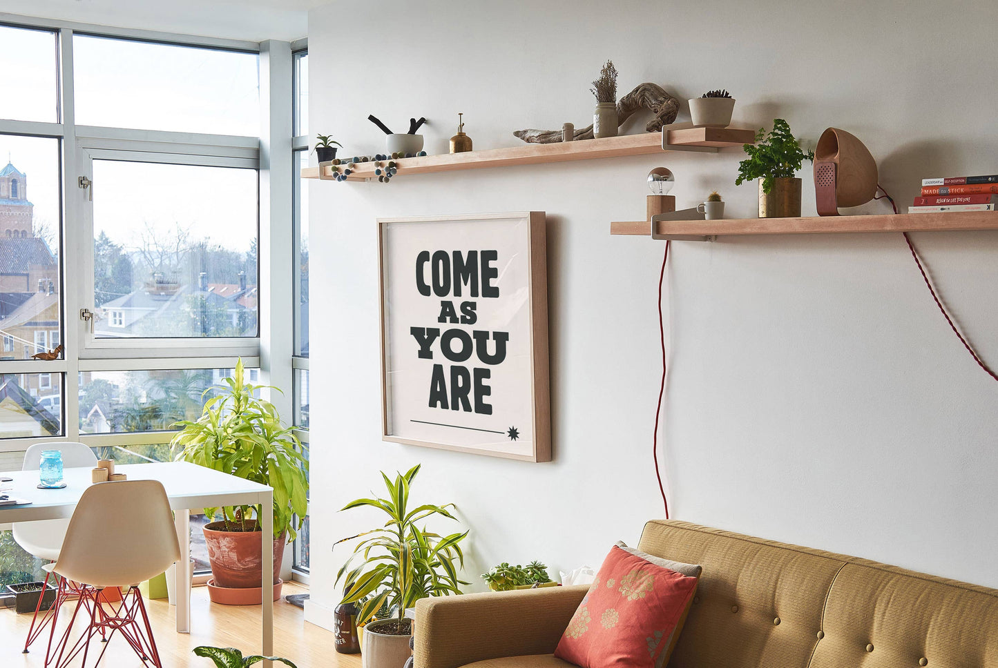 Modern living room with a 'Come as You Are' framed quote, plants, and a sofa.