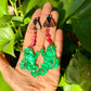 Hand holding two green frog earrings with pink ribbons against a leafy green background