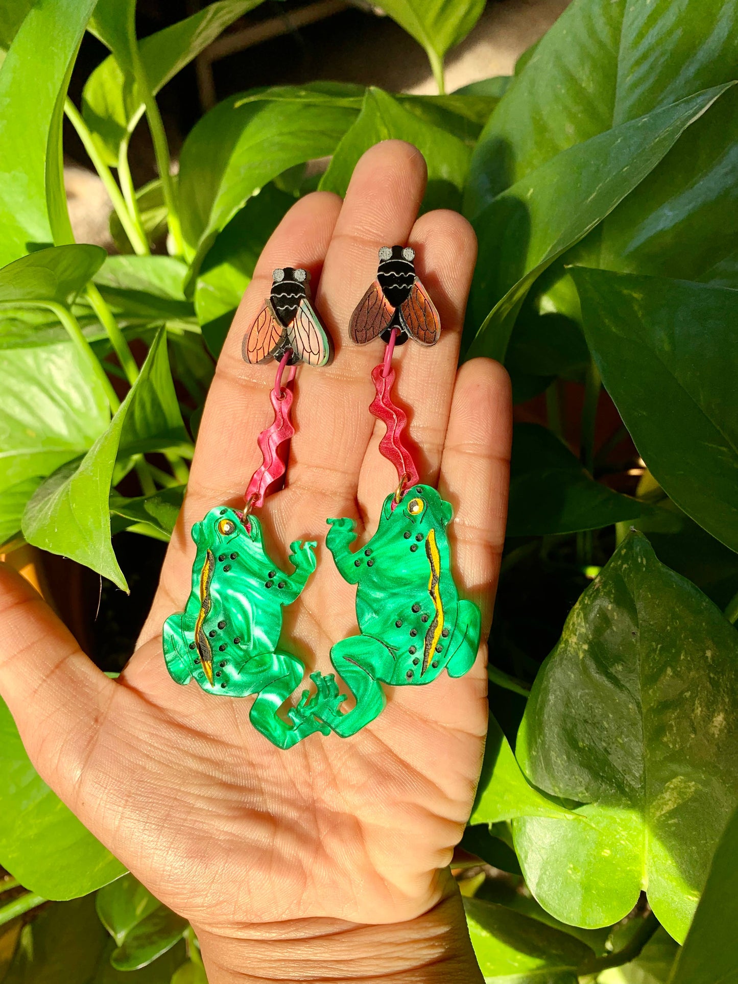 Hand holding two green frog earrings with pink ribbons against a leafy green background