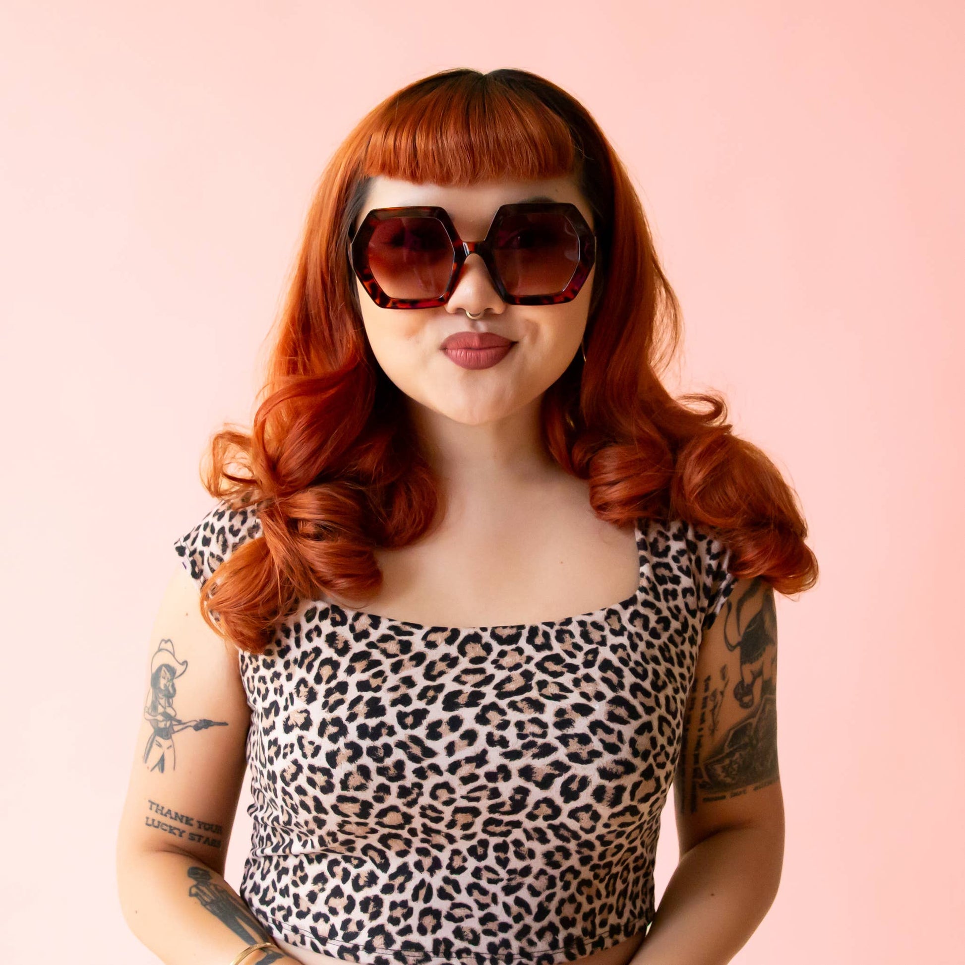 Woman with red hair and sunglasses wearing a leopard print top on a pink background