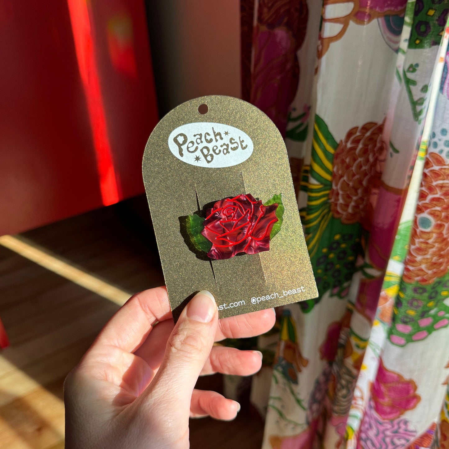 Hand holding a rose hair clip with 'Peach Beast' branding against a colorful background