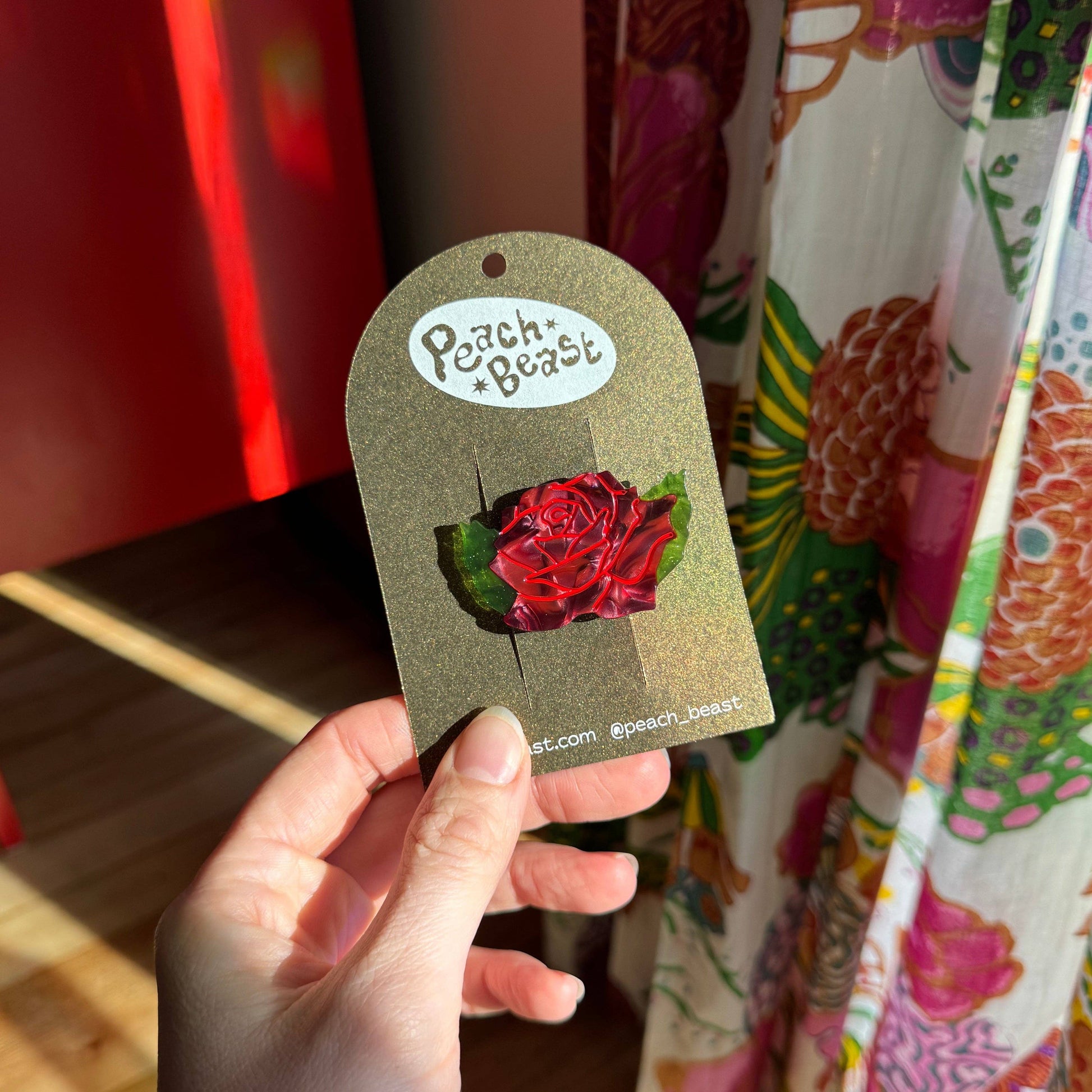 Hand holding a rose hair clip with 'Peach Beast' branding against a colorful background