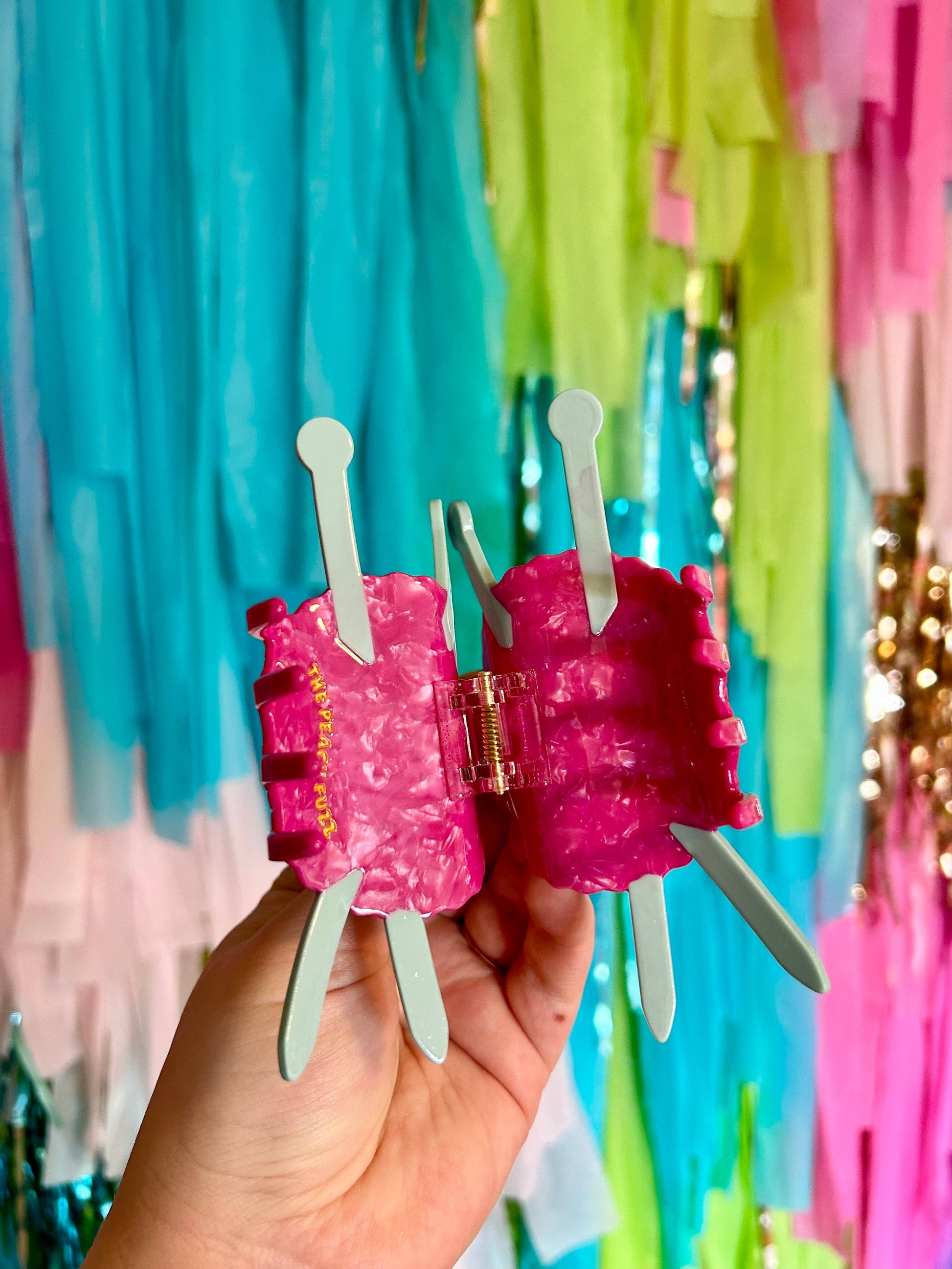 Pink knitting yarn hair clips held by a hand with a colorful fringe background