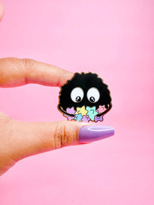 Black soot sprite with white eyes and colorful polka dots held by a hand against a pink background