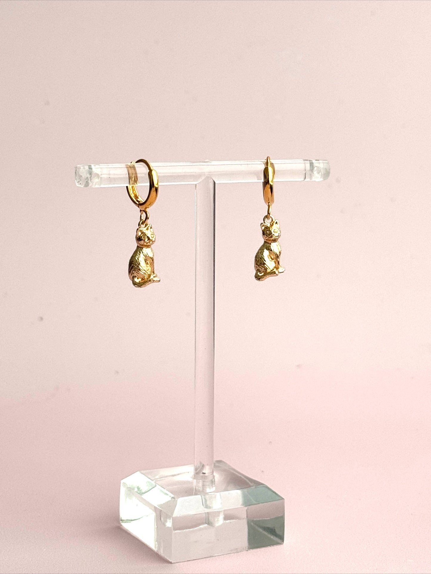Gold cat hoop earrings on a clear stand against a pink background