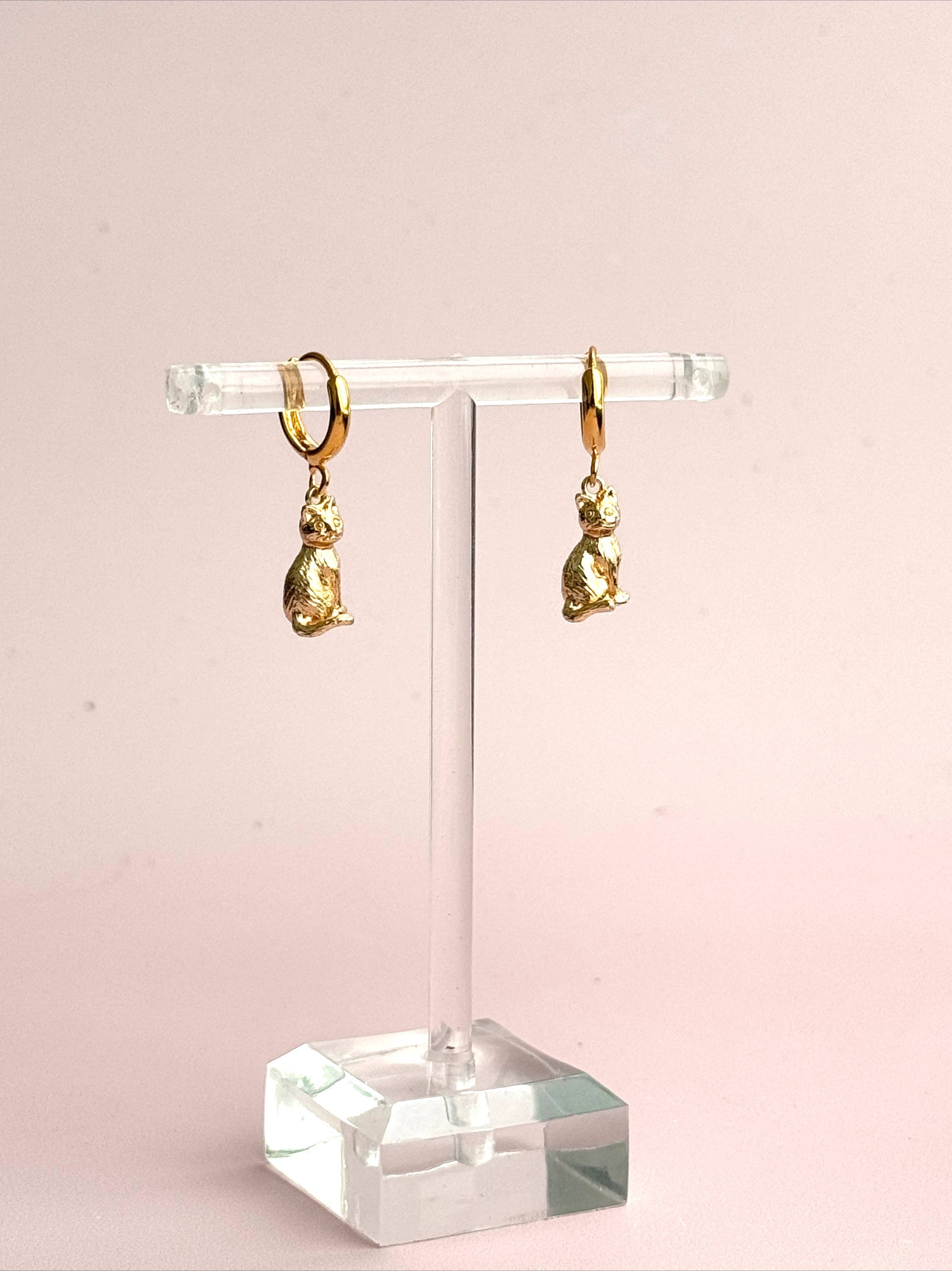 Gold cat hoop earrings on a clear stand against a pink background