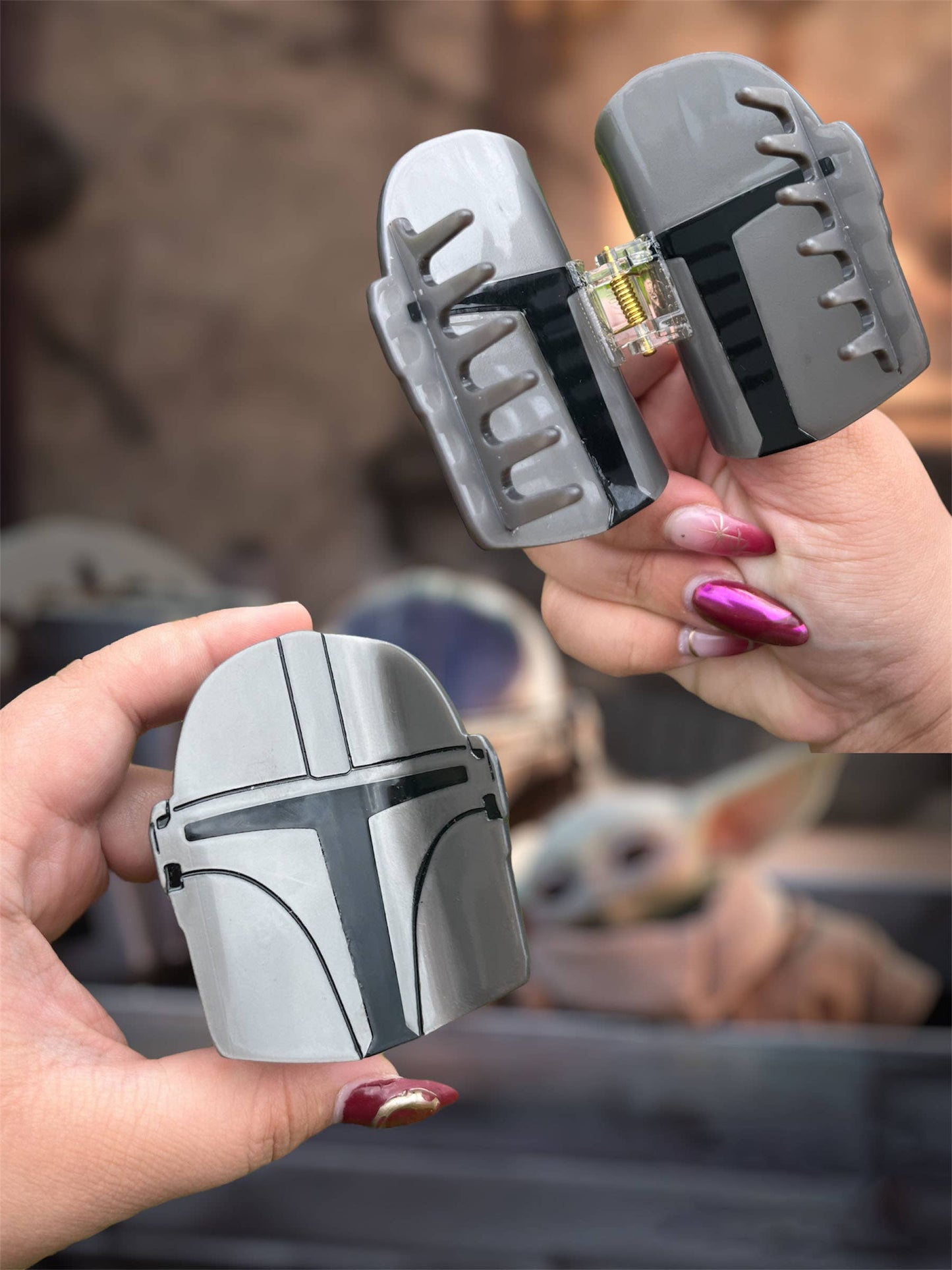 Two hands holding gray and silver Mandalorian helmet-shaped hair claw clips