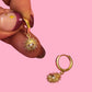 Gold hoop earrings with sunflower designs held by a hand against a pink background