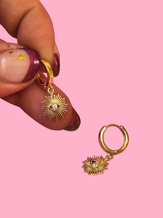 Gold hoop earrings with sunflower designs held by a hand against a pink background