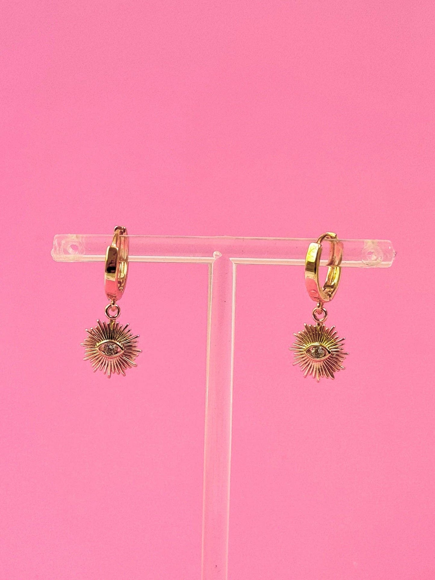 Gold hoop earrings with sunray shaped charms on a clear stand against a pink background