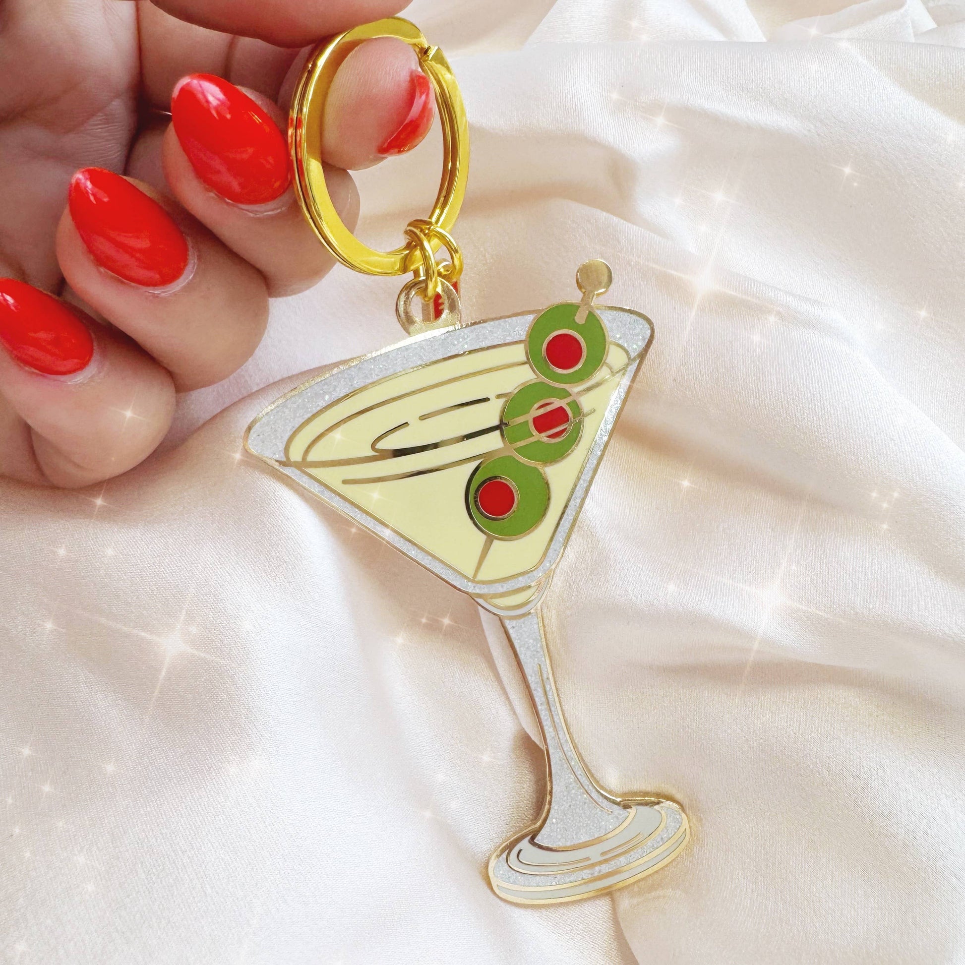 Keychain shaped like a martini glass with olives held by a hand with red nail polish on a light background.