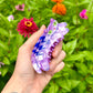 Hand holding a colorful plastic insect-shaped toy against a background of green foliage and flowers.