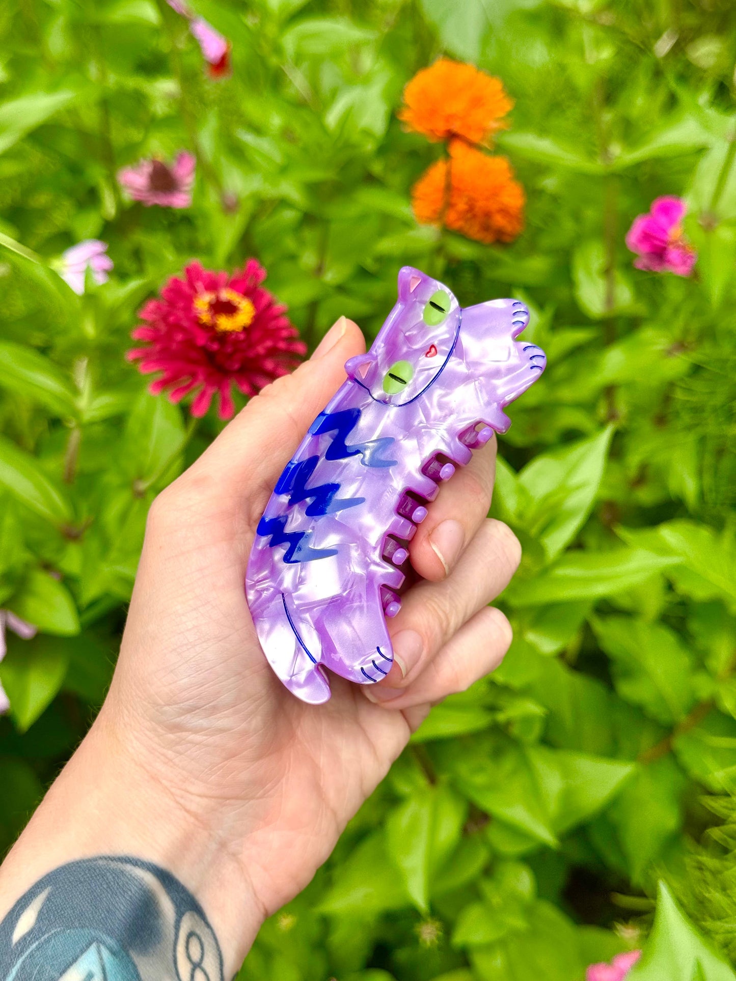 Hand holding a colorful plastic insect-shaped toy against a background of green foliage and flowers.