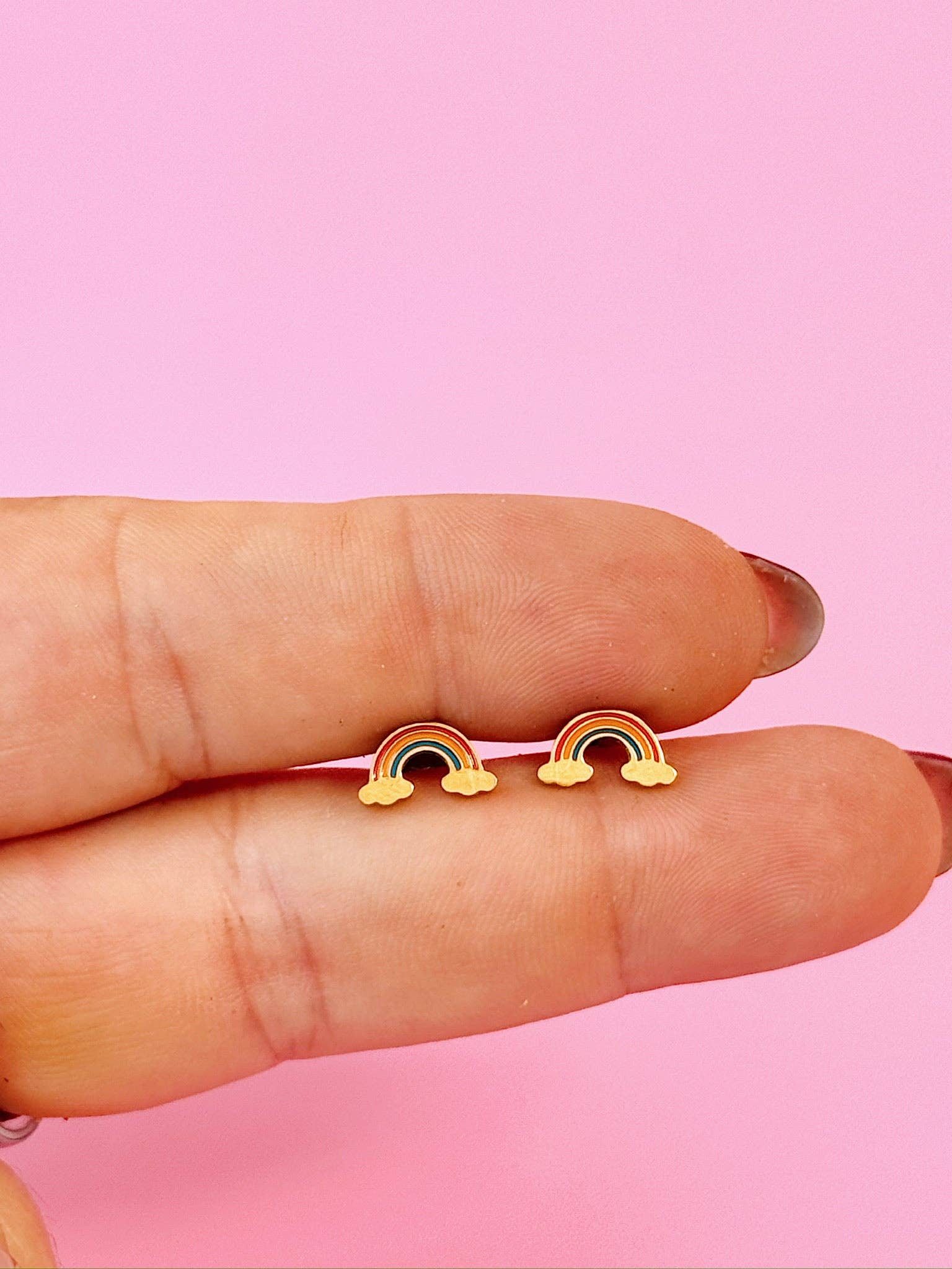 Rainbow-shaped earrings held between fingers against a pink background