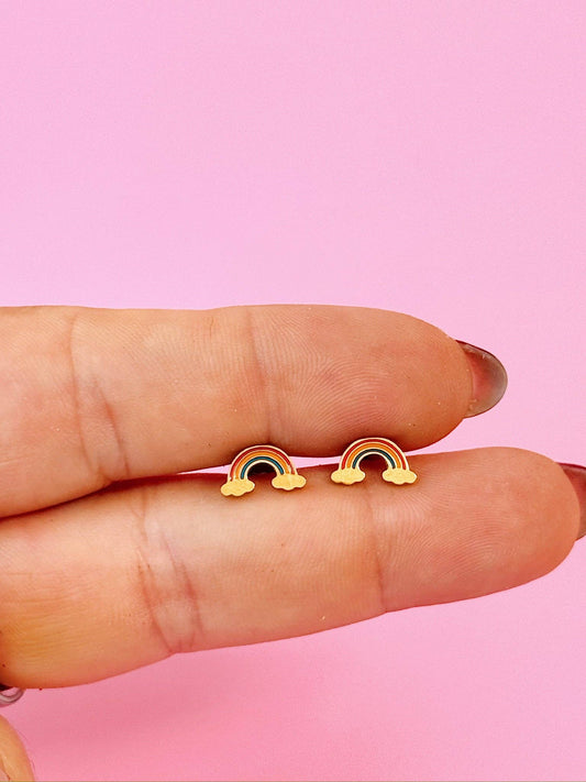 Rainbow-shaped earrings held between fingers against a pink background
