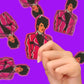 Hand holding a card with a stylized illustration of Prince in a red jacket on a purple background.