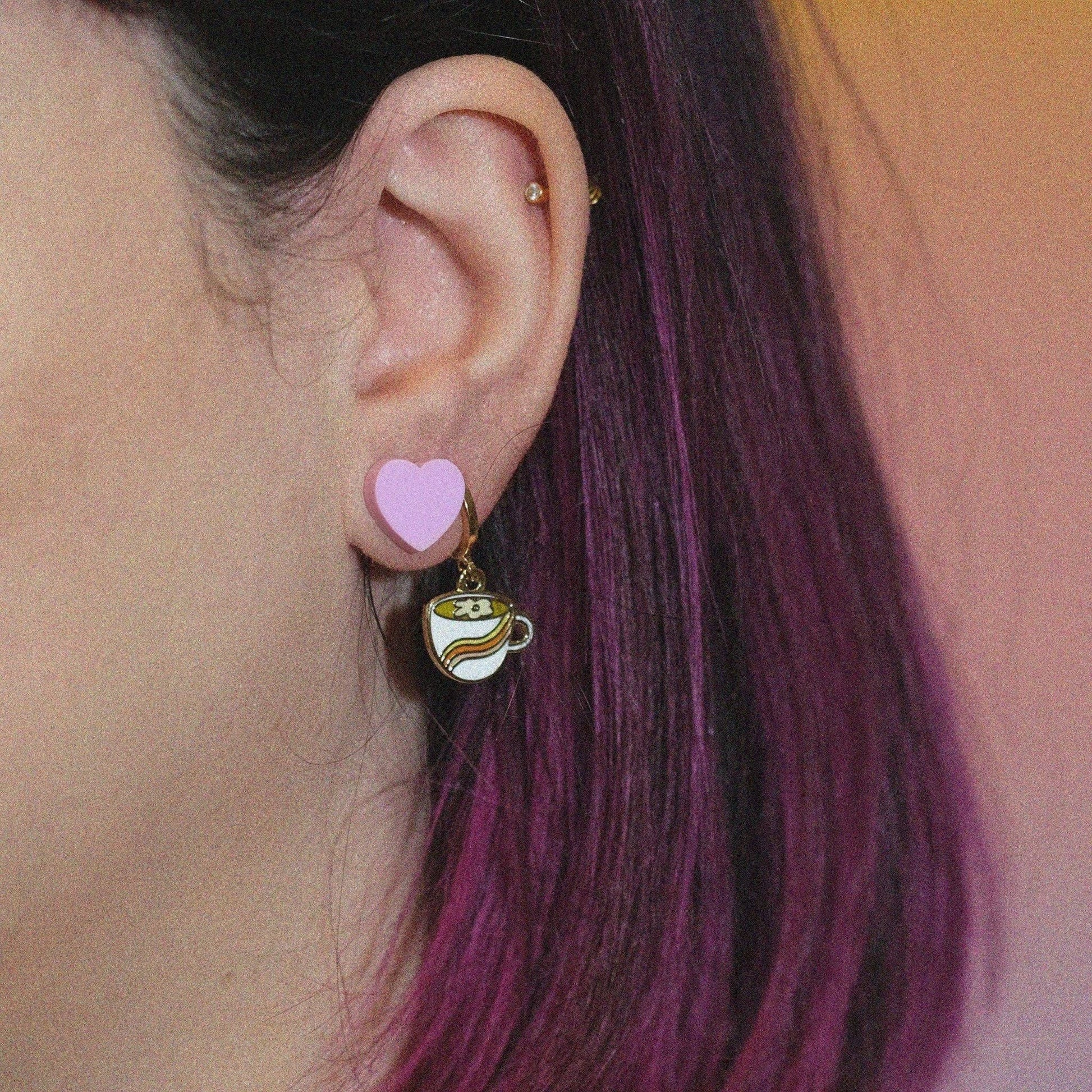 Close-up of an ear with a pink heart earring and a coffee cup earring against a blurred background.