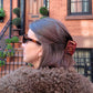 Person with gray hair wearing a Pride and Prejudice book claw clip and a fur-lined coat in front of a brick building.