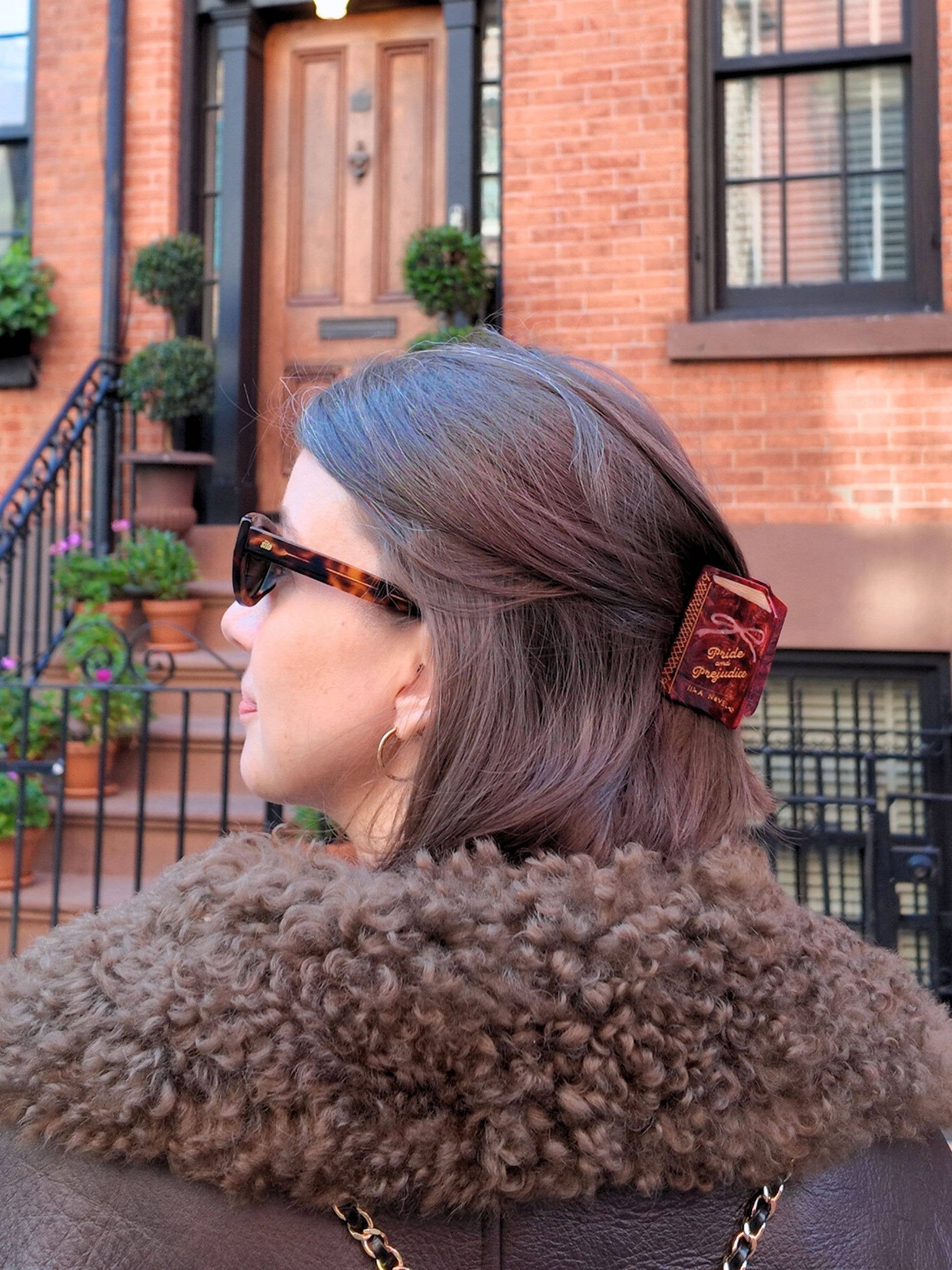 Person with gray hair wearing a Pride and Prejudice book claw clip and a fur-lined coat in front of a brick building.