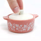 Pink pyrex ceramic container with floral patterns and a white lid being held by a hand on a white background