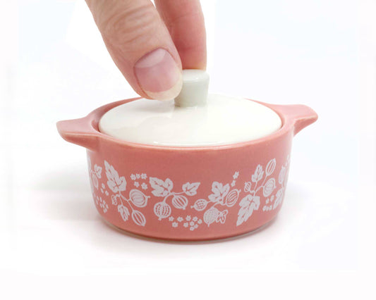 Pink pyrex ceramic container with floral patterns and a white lid being held by a hand on a white background