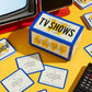 Card game box titled 'Poorly Explained TV Shows' on a yellow surface with cards around it.