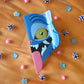 Blue tiger hair claw with a pink tongue on an orange background with colorful shapes