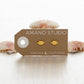 Gold lip-shaped earrings on a 'Amano Studio' branded card with seashells in the background.