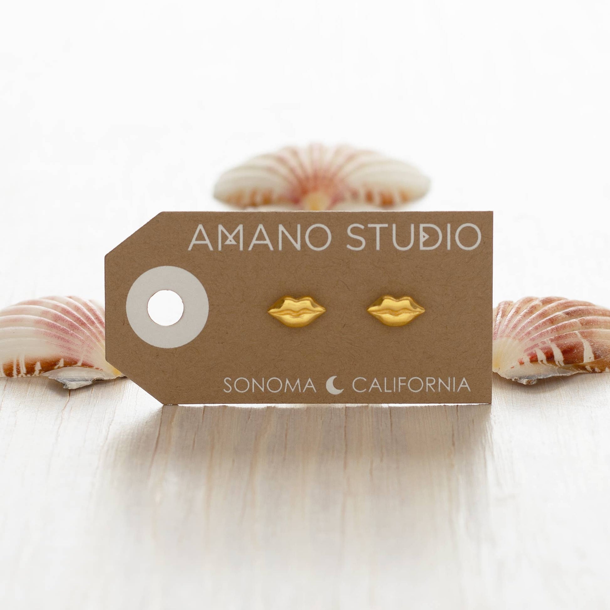 Gold lip-shaped earrings on a 'Amano Studio' branded card with seashells in the background.