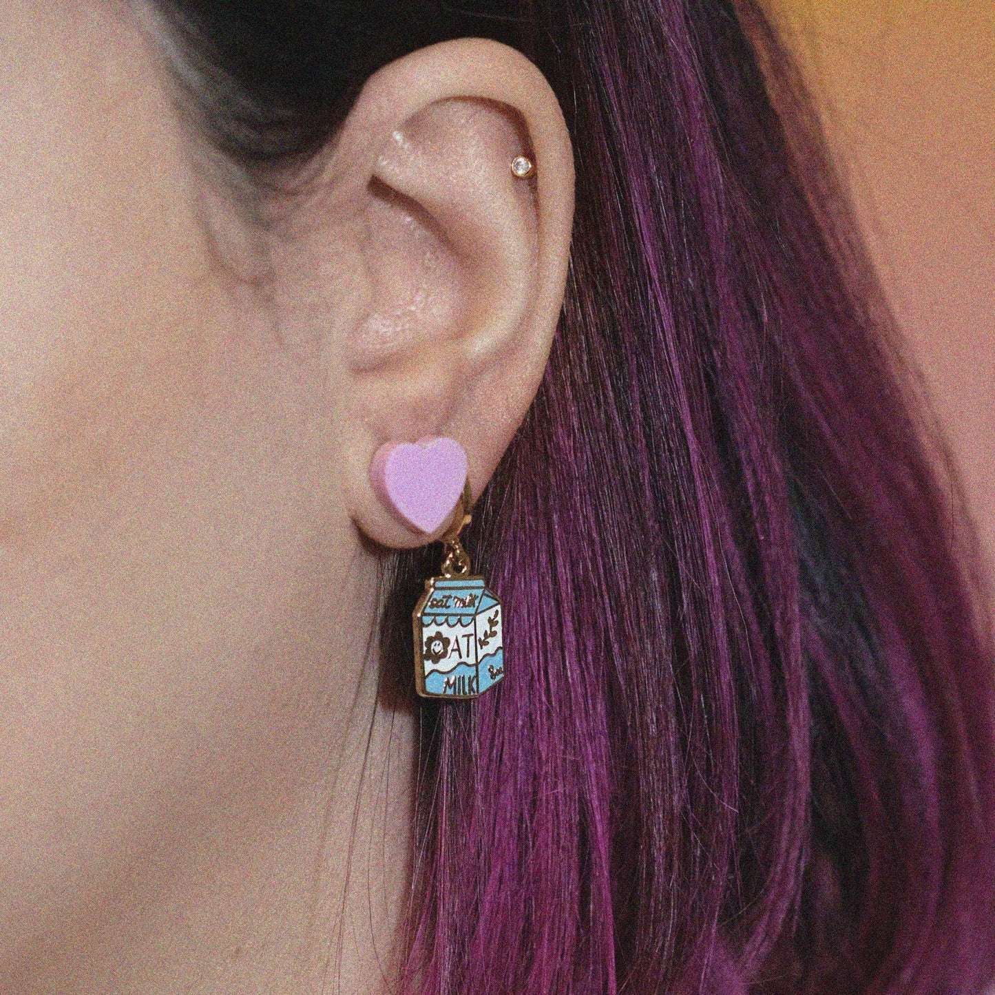 Close-up of an ear with a heart-shaped earring and a colorful oat milk earring.