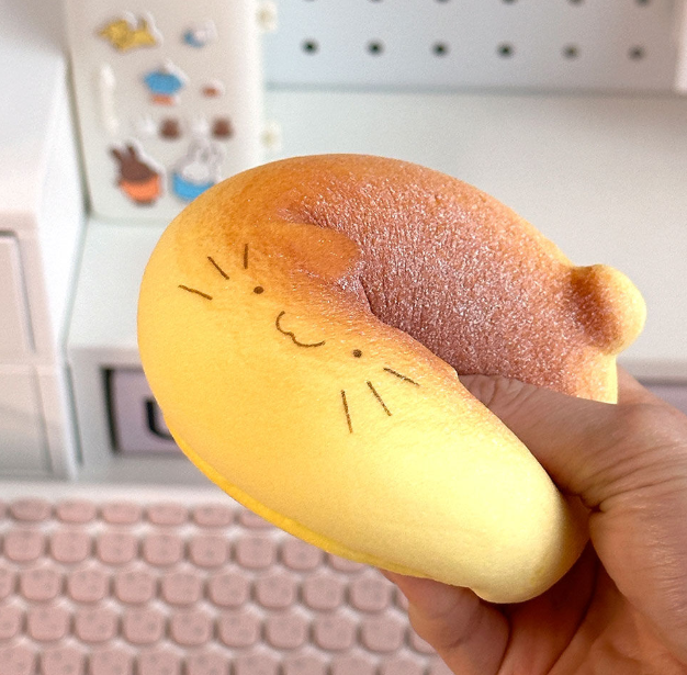 Hand holding a squishy toy shaped like a cat made of bread with a textured surface, blurred office background.