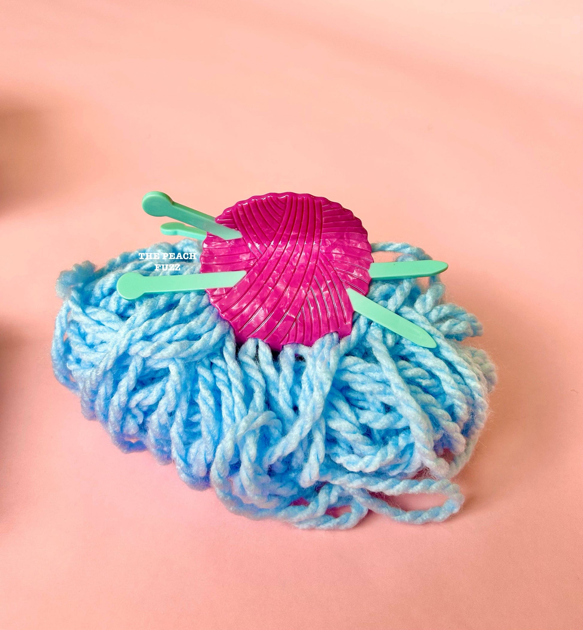Blue yarn ball with a pink knitting yarn claw clip and green sticks on a pink background