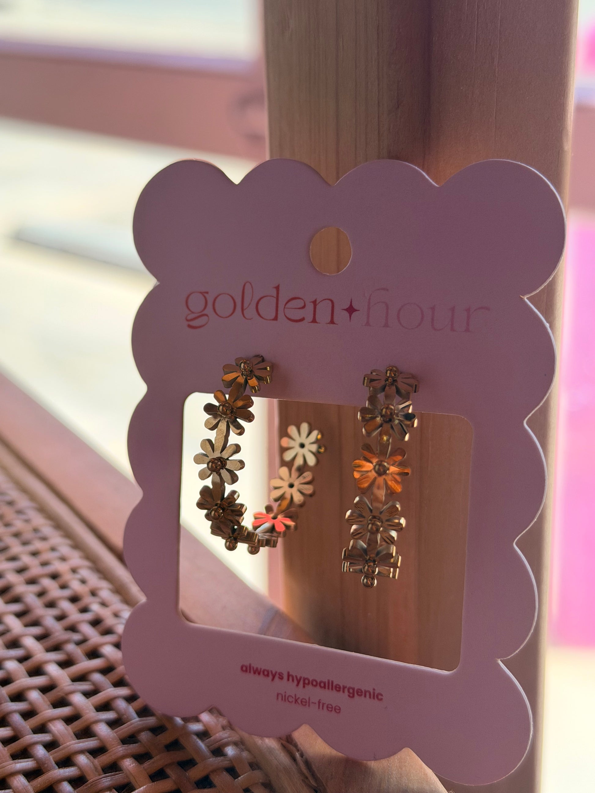 A pair of flower-shaped gold hoop earrings displayed on a pink scalloped card labeled 'golden hour', resting on a woven surface with a wooden post in the background.