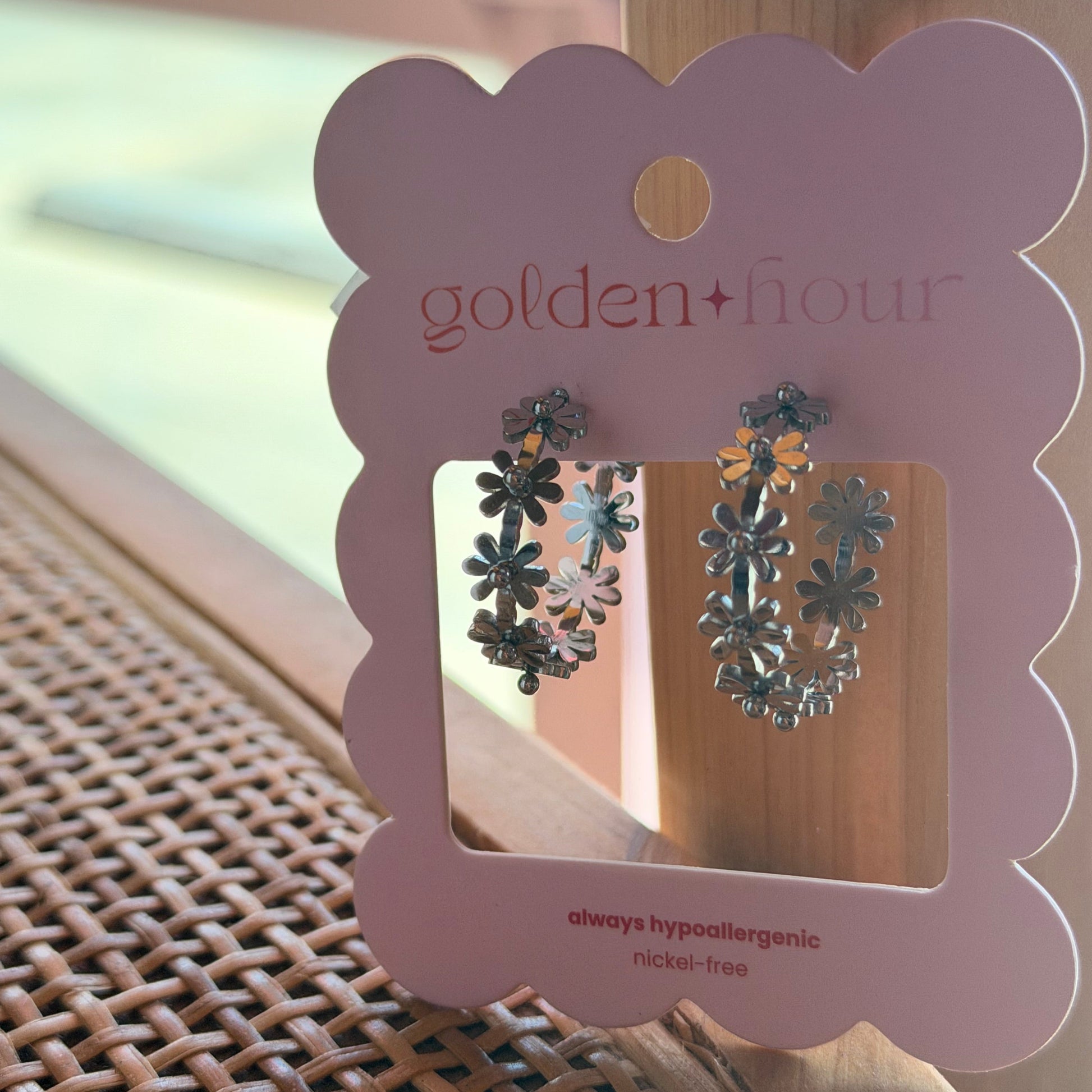 Silver floral hoop earrings displayed on a pink scalloped card labeled 'golden hour', resting on a woven wicker surface near a wooden post.