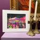 Framed artwork of a colorful building on a purple surface with candles and a painting in the background.