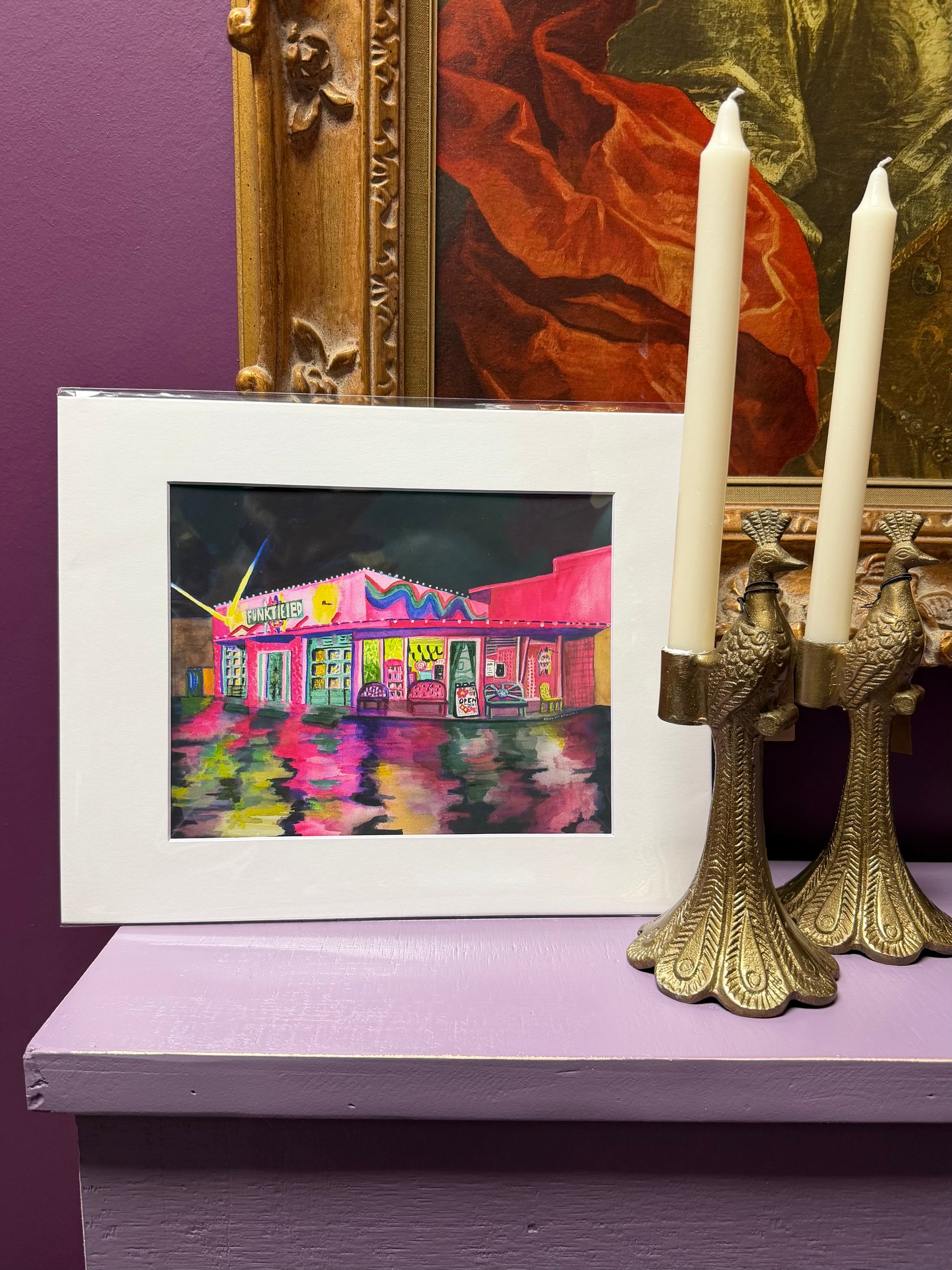 Framed artwork of a colorful building on a purple surface with candles and a painting in the background.
