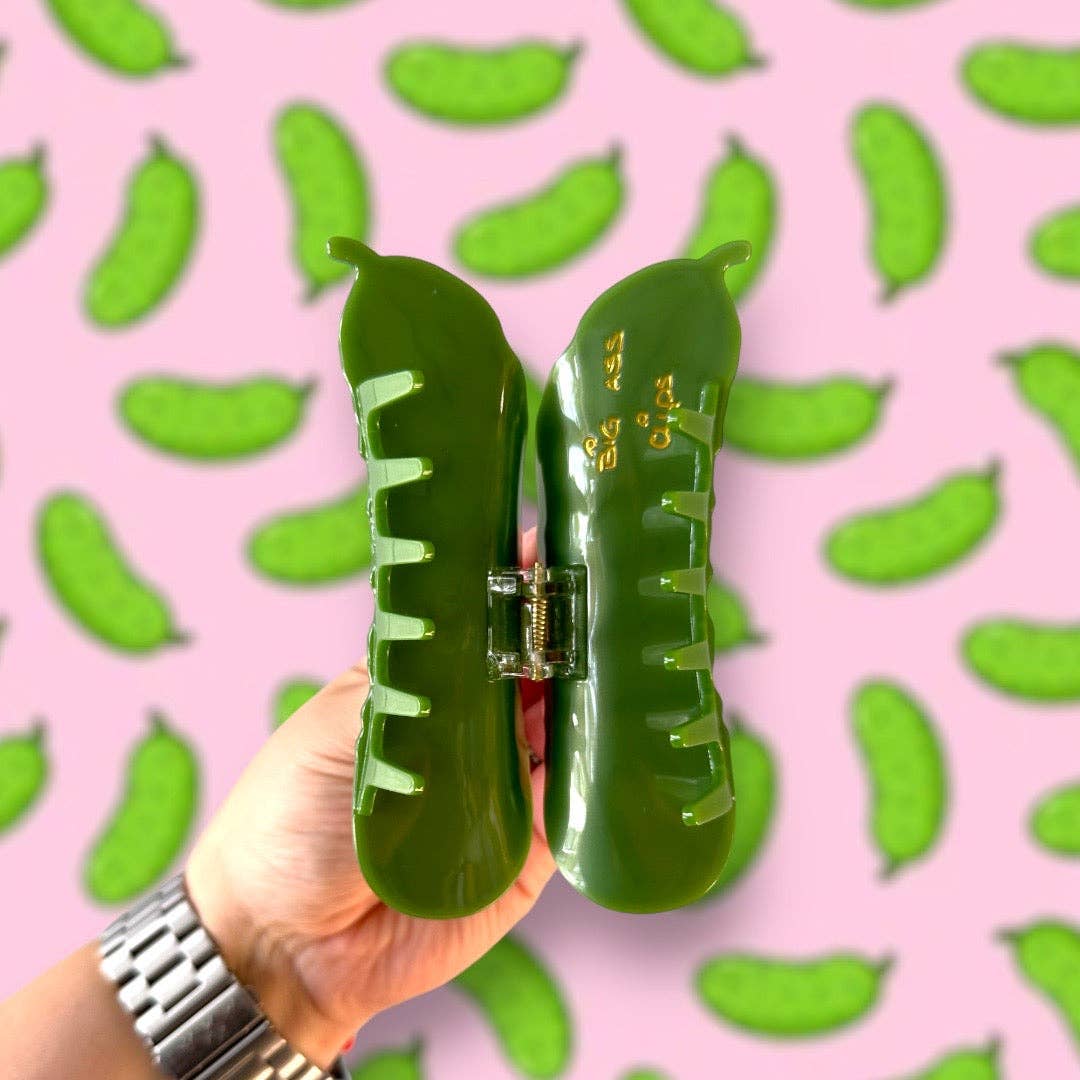 Green pickle hair claw held by a hand against a pink background with green leaf patterns.