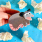 Hand holding a possum-shaped Hair claw clip against a blue background with crumpled white paper.