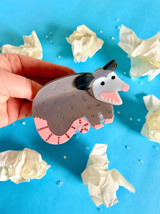 Hand holding a possum-shaped Hair claw clip against a blue background with crumpled white paper.