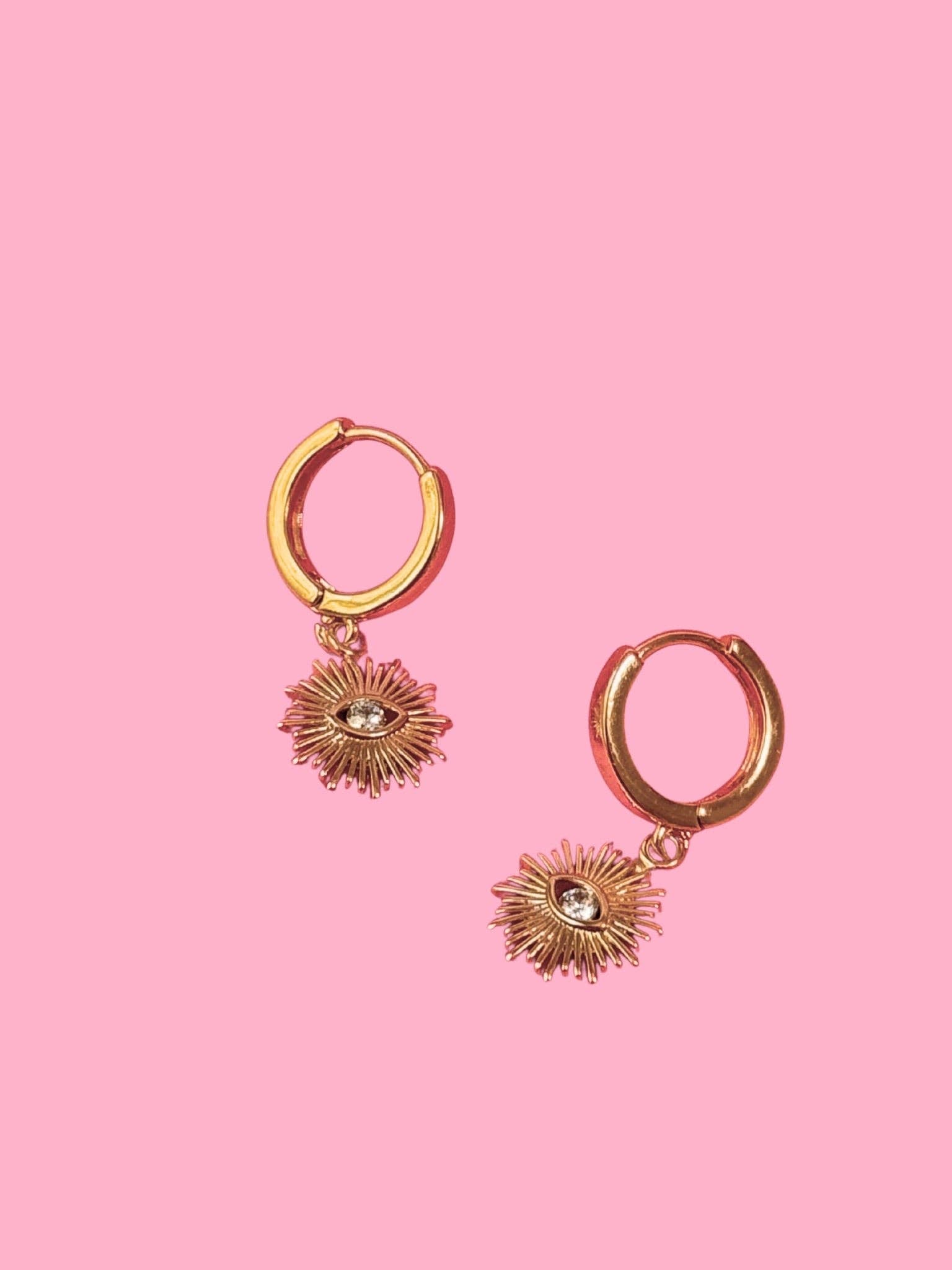 Gold hoop earrings with sunray-shaped charms on a pink background