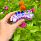 Hand holding a purple and blue cat-shaped object in front of a garden with flowers.