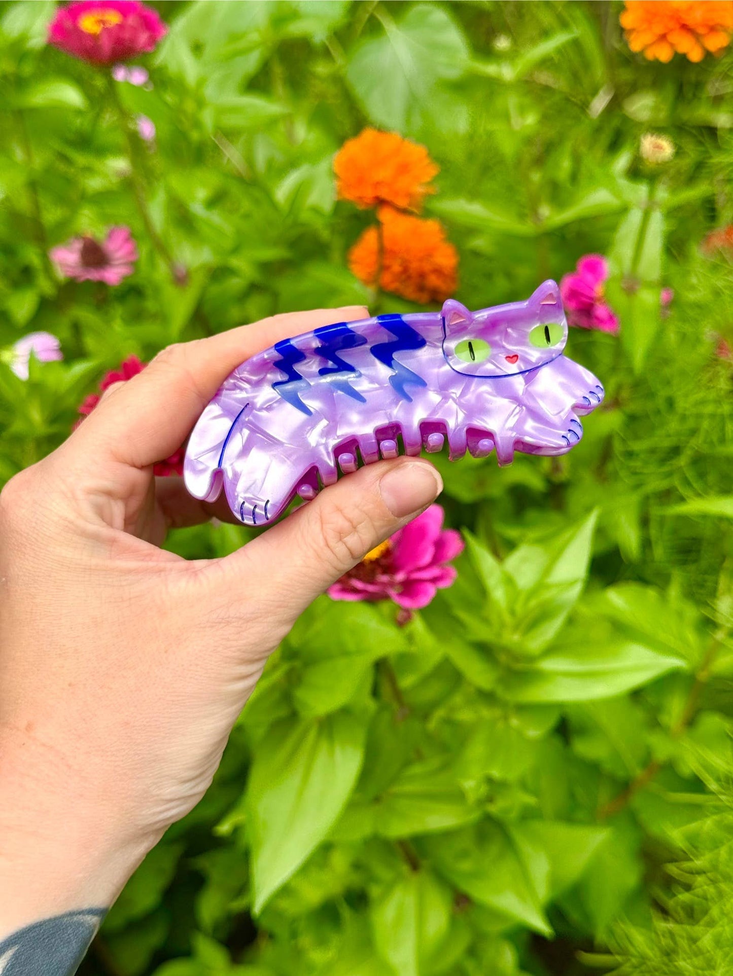 Hand holding a purple and blue cat-shaped object in front of a garden with flowers.
