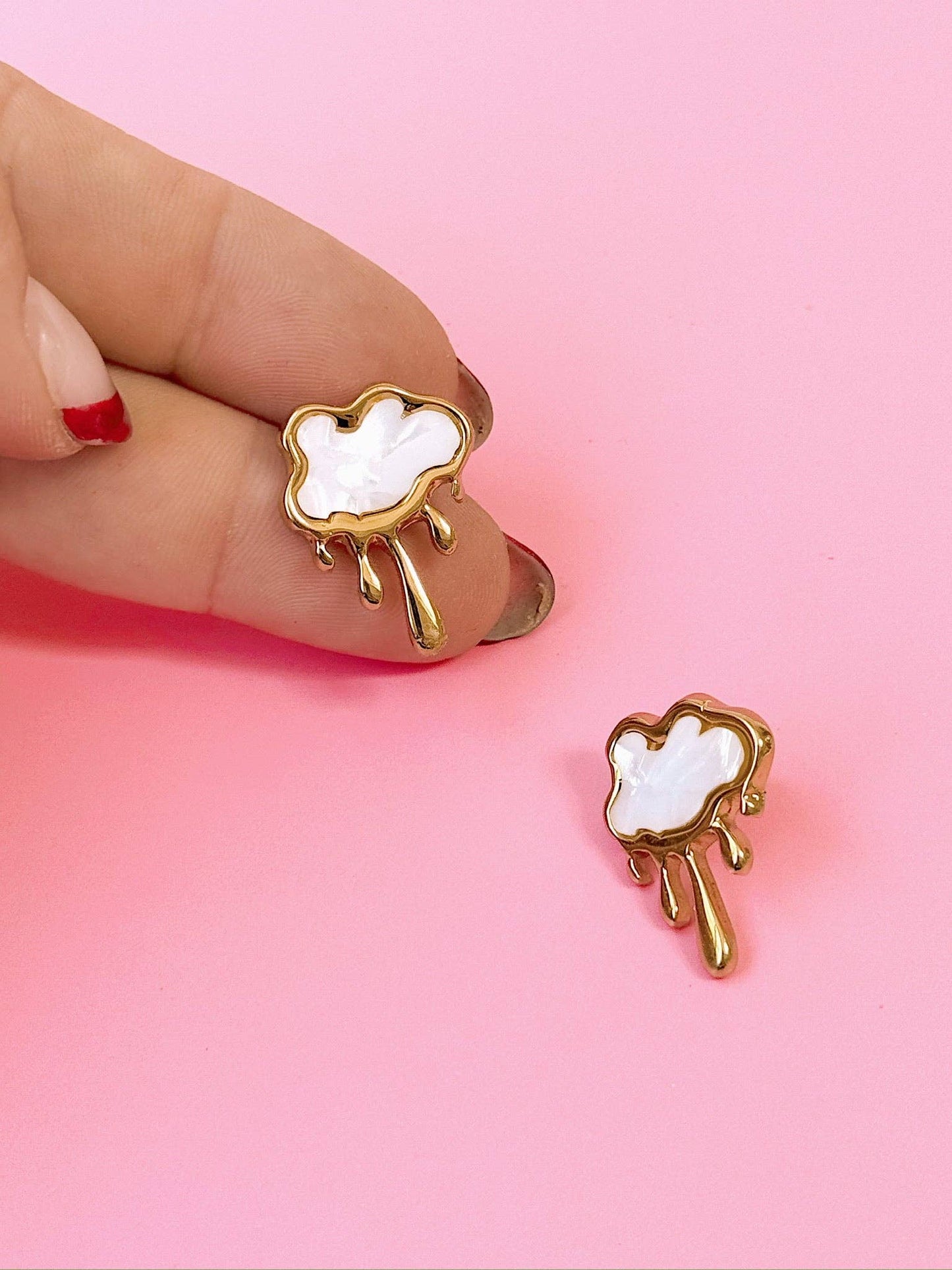 Cloud-shaped earrings with gold accents held by a hand on a pink background