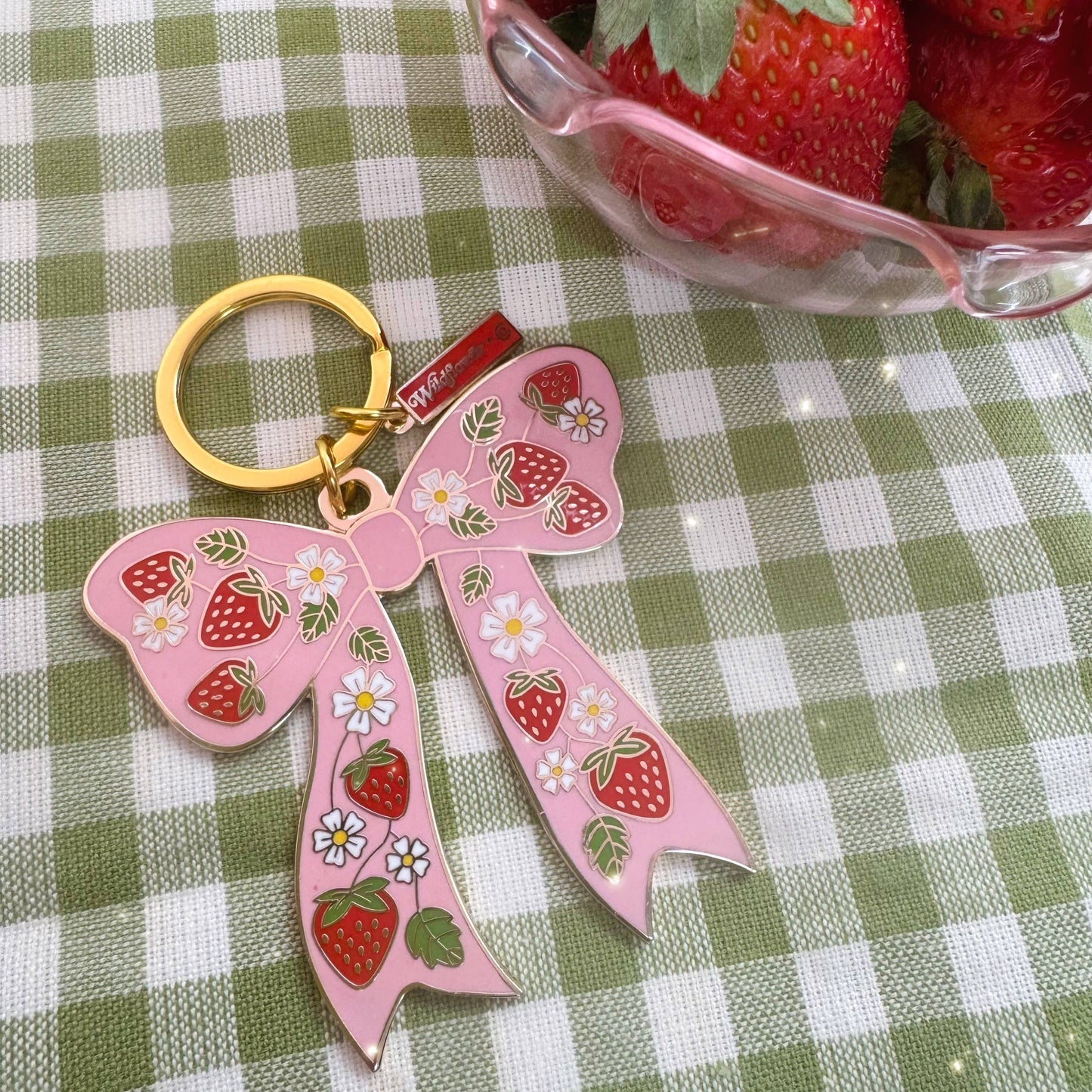 Pink bow keychain with strawberry design on a green and white checkered tablecloth.