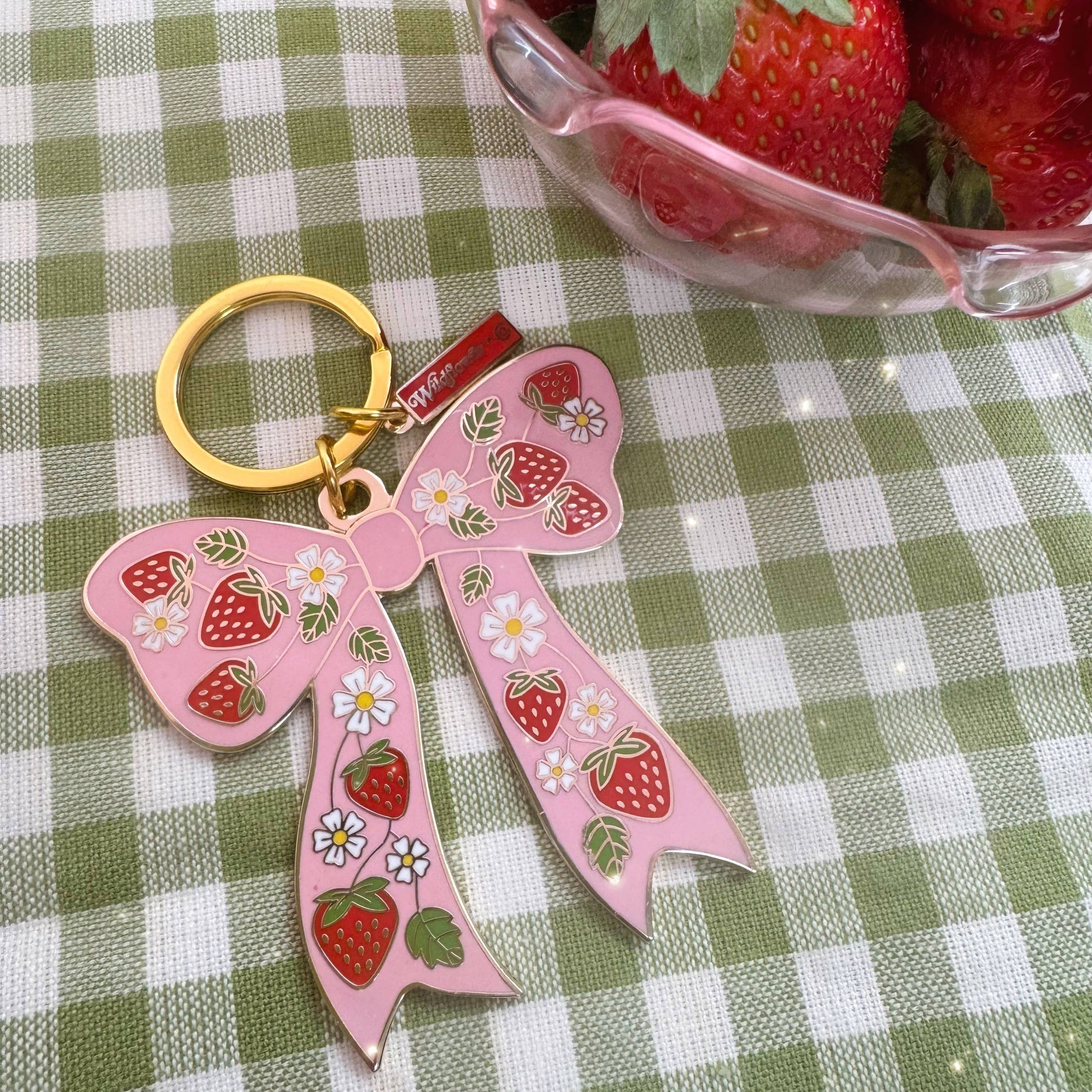 Pink bow keychain with strawberry design on a green and white checkered tablecloth.