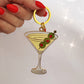 Keychain shaped like a martini glass with olives held by a hand with red nail polish on a white background.
