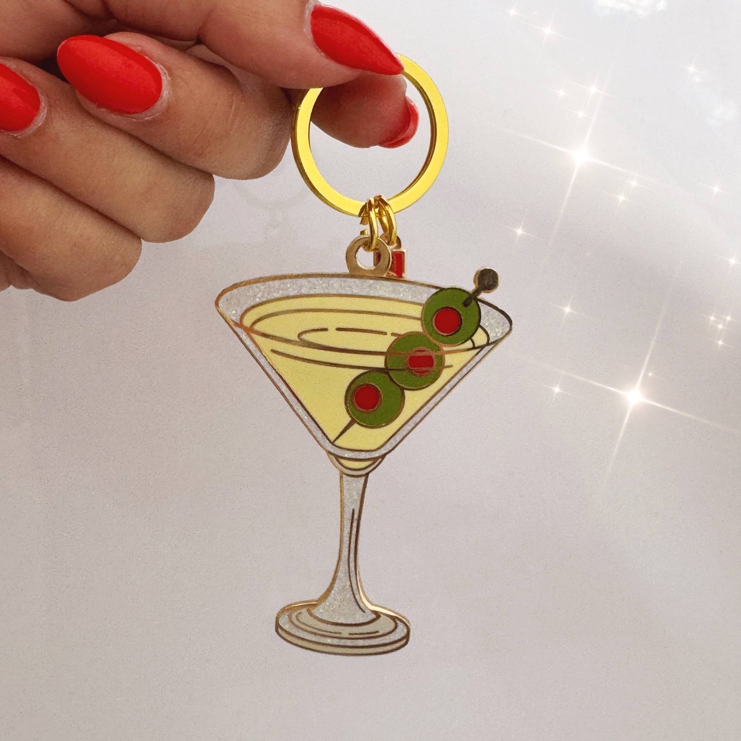 Keychain shaped like a martini glass with olives held by a hand with red nail polish on a white background.