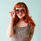 Woman with red hair wearing sunglasses and a leopard print top against a light blue background