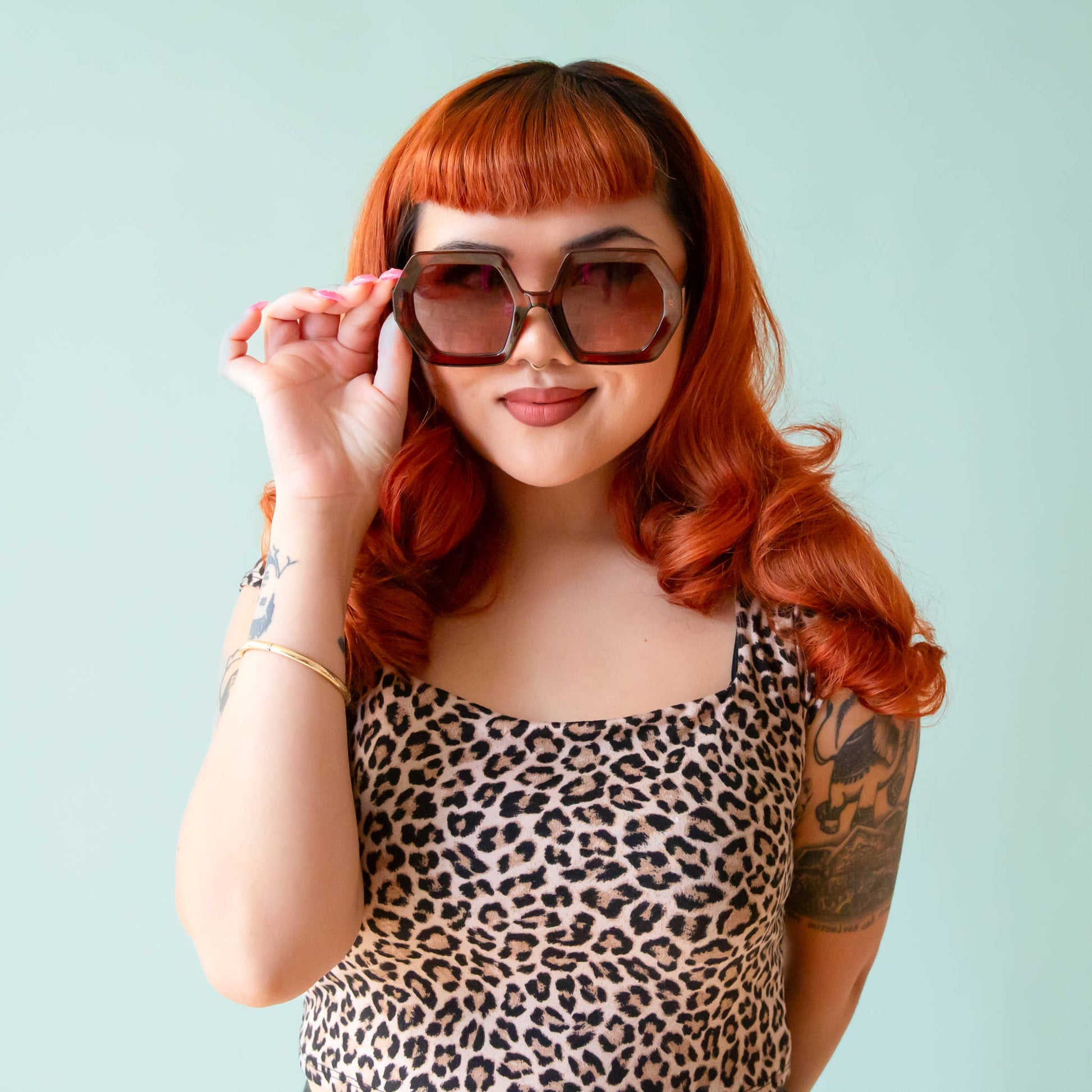 Woman with red hair wearing sunglasses and a leopard print top against a light blue background